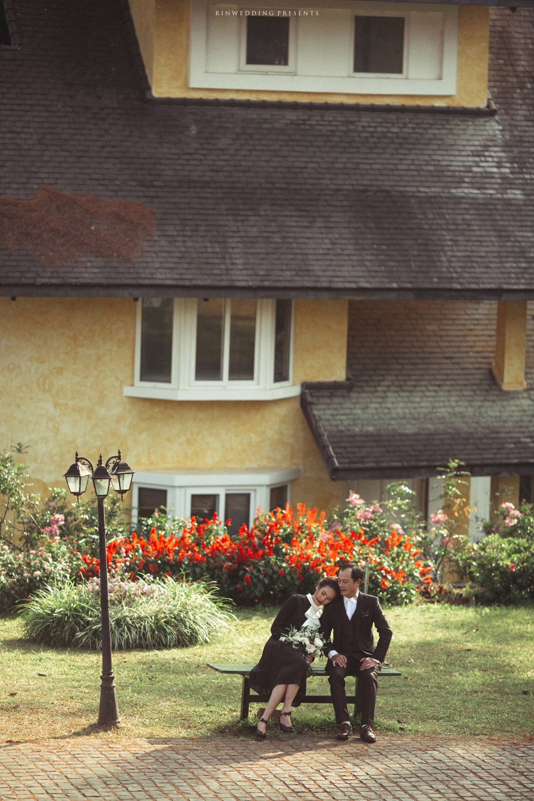A bride and groom sit beside flowers before a pitched-roof villa.