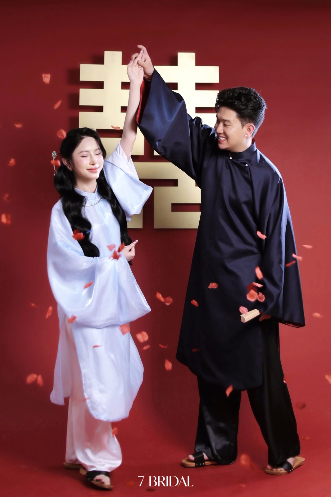 A couple in áo dài holds hands aloft before a golden double-happiness symbol.