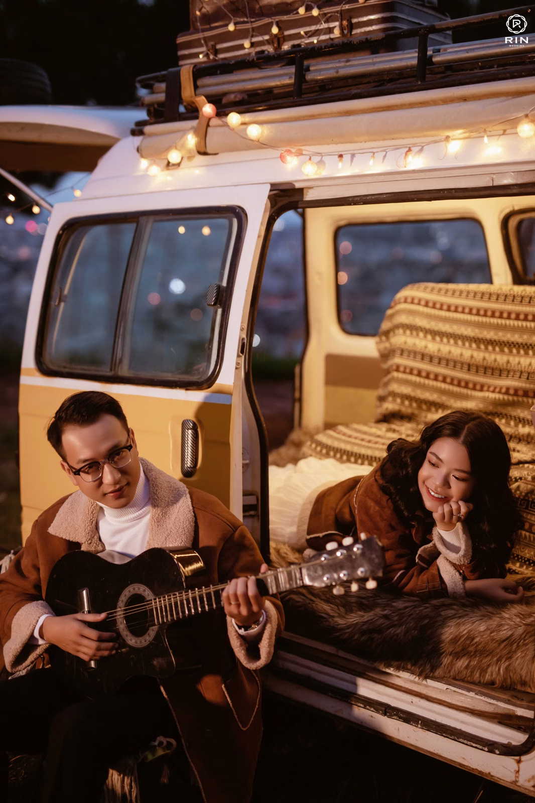 A groom plays guitar beside the bride at the doorway of a camper decorated with string lights.