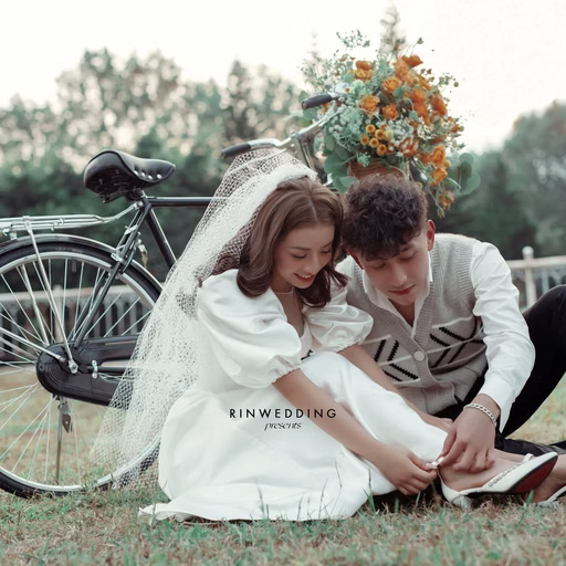 The bride and groom sit on the grass beside a bicycle and orange flowers.