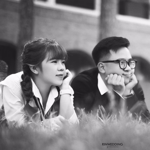 Black-and-white photograph of the couple lying on grass before Cao Đẳng Sư Phạm Đà Lạt.