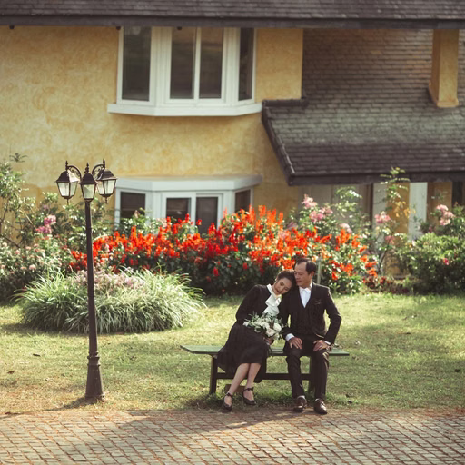 A bride and groom sit beside flowers before a pitched-roof villa.
