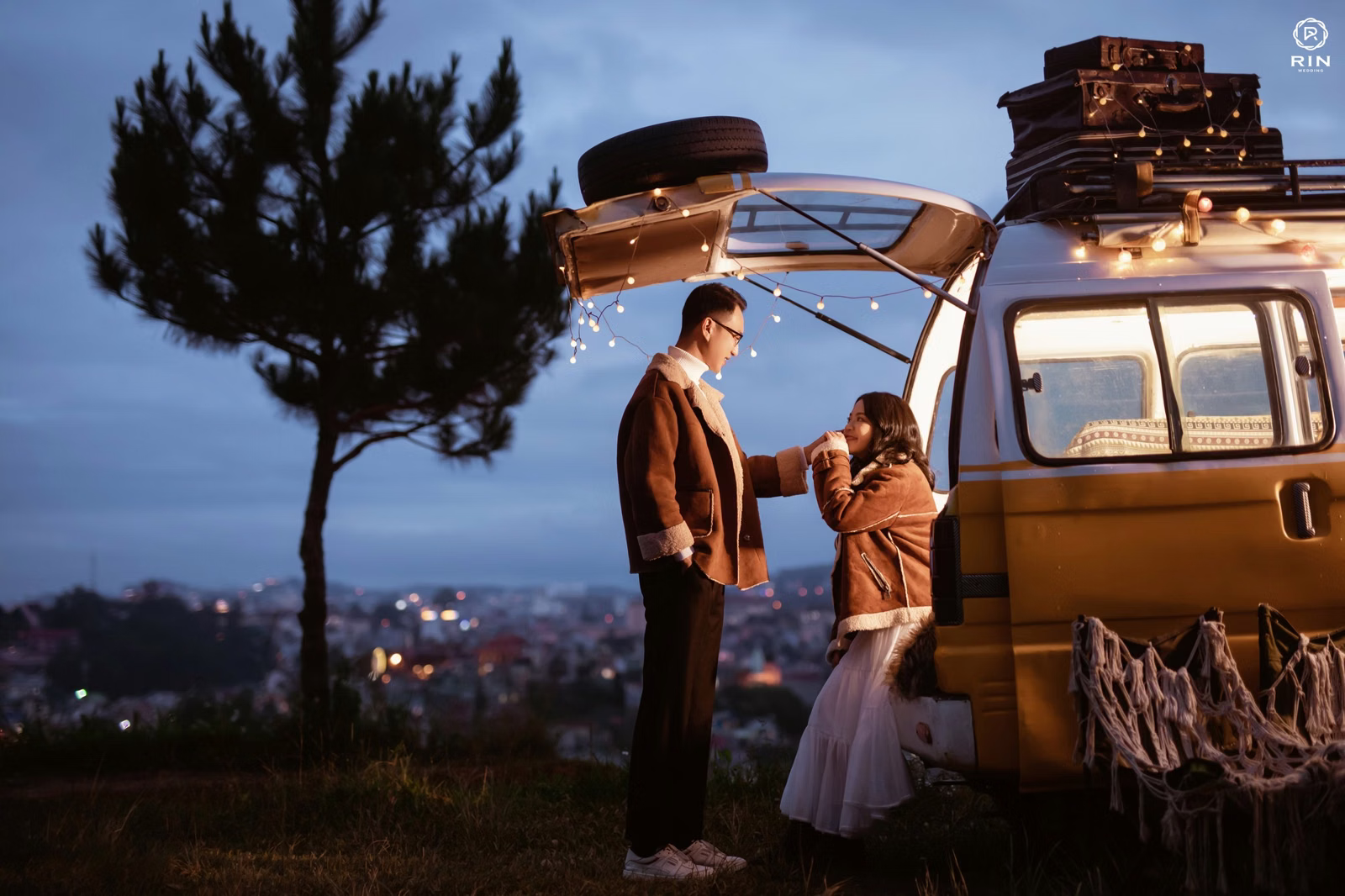A couple chatting beside a camper vehicle, with city lights in the distance.
