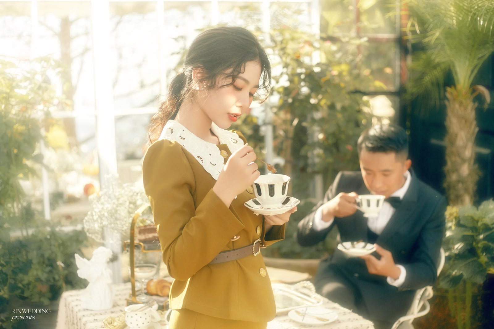 A woman in a yellow dress raising a teacup beside a man in a suit in a sunlit café.