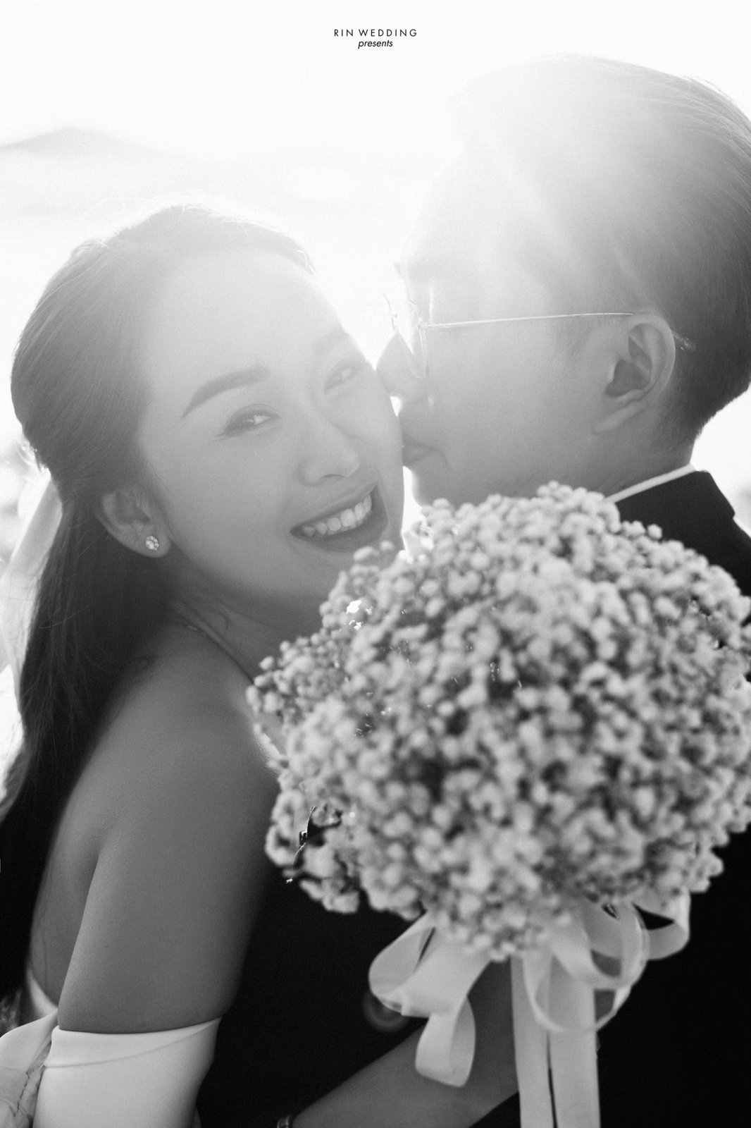 A black-and-white photograph of the groom kissing the bride's cheek in sunshine at Đồi Thông Đà Lạt