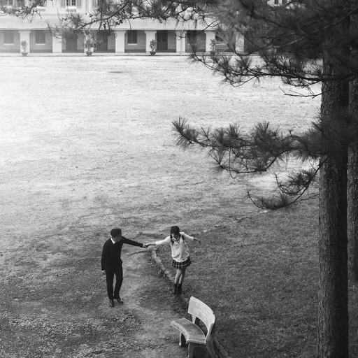 Black-and-white photograph of the couple holding hands beneath pine trees at Cao Đẳng Sư Phạm Đà Lạt.