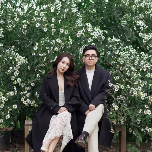 The couple sits close on a wooden bench before a white floral arch in Đà Lạt.