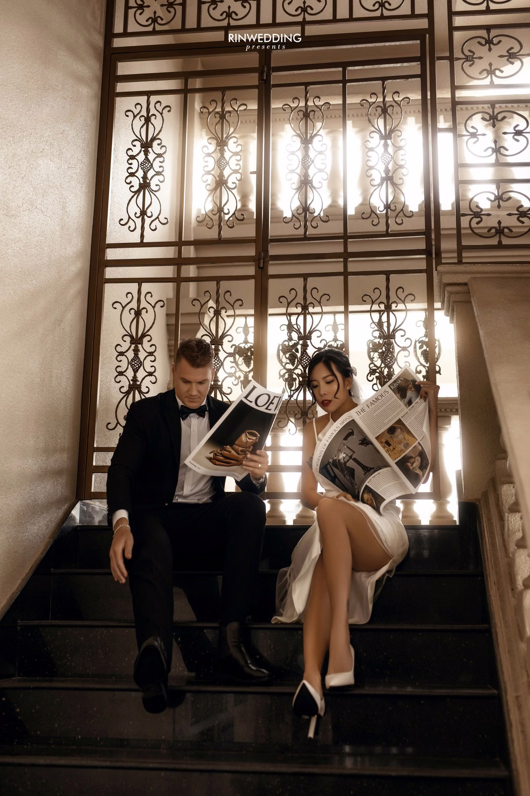 The bride and groom reading newspapers on steps before Don Bosco's decorative iron doors.