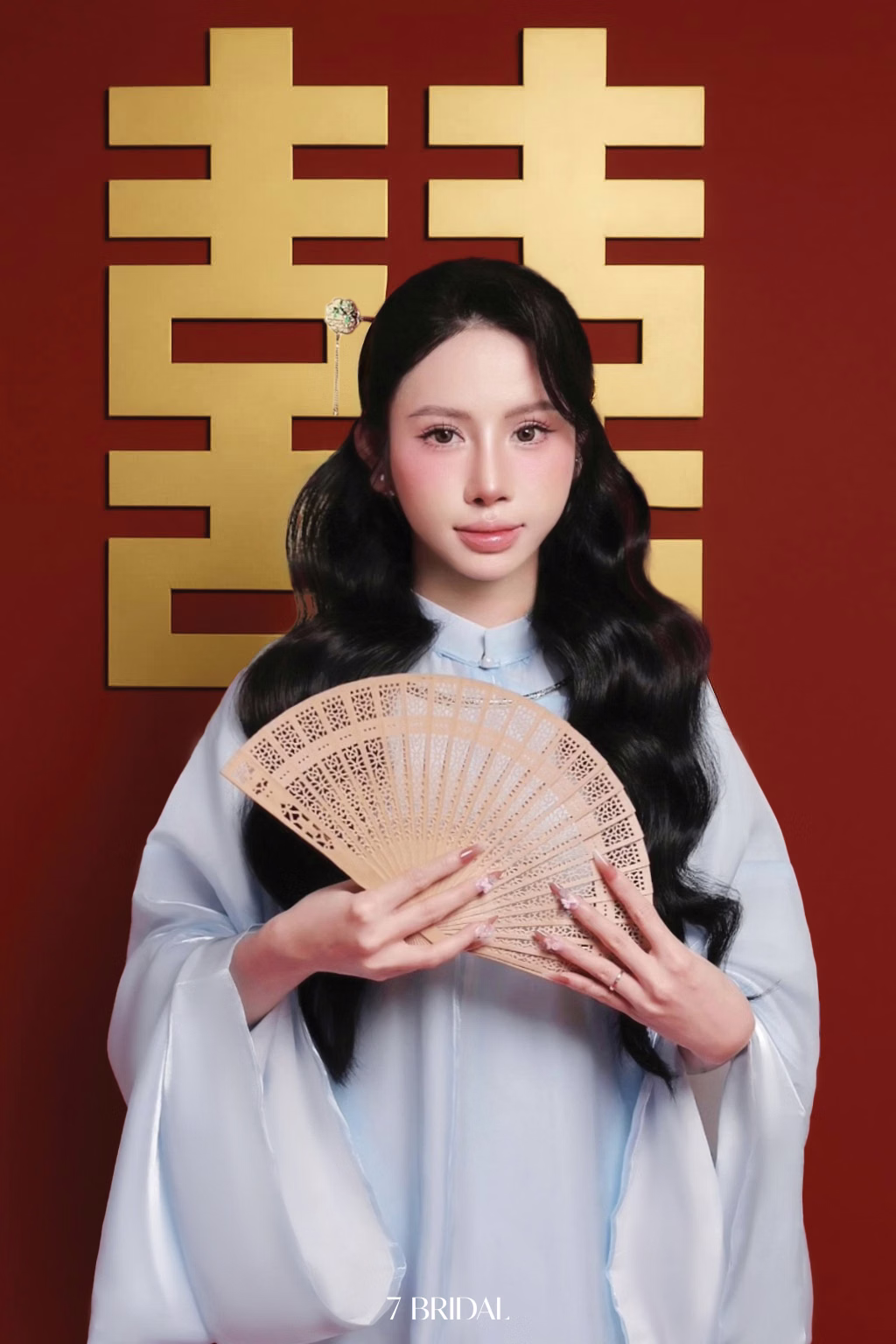 The bride in a pale blue áo dài holding a fan before a red backdrop with the double-happiness symbol.