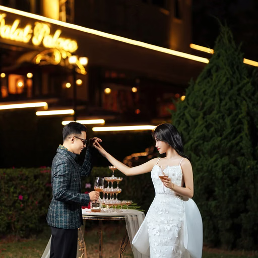 The bride touches the groom's sunglasses before an illuminated hotel.
