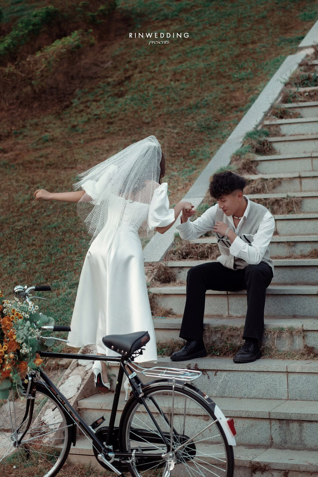 The bride holds the groom's hand while seated on steps beside a flower-decorated bicycle.