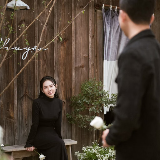 A man holds flowers and looks at his girlfriend seated at a table outside a wooden café in Đà Lạt.