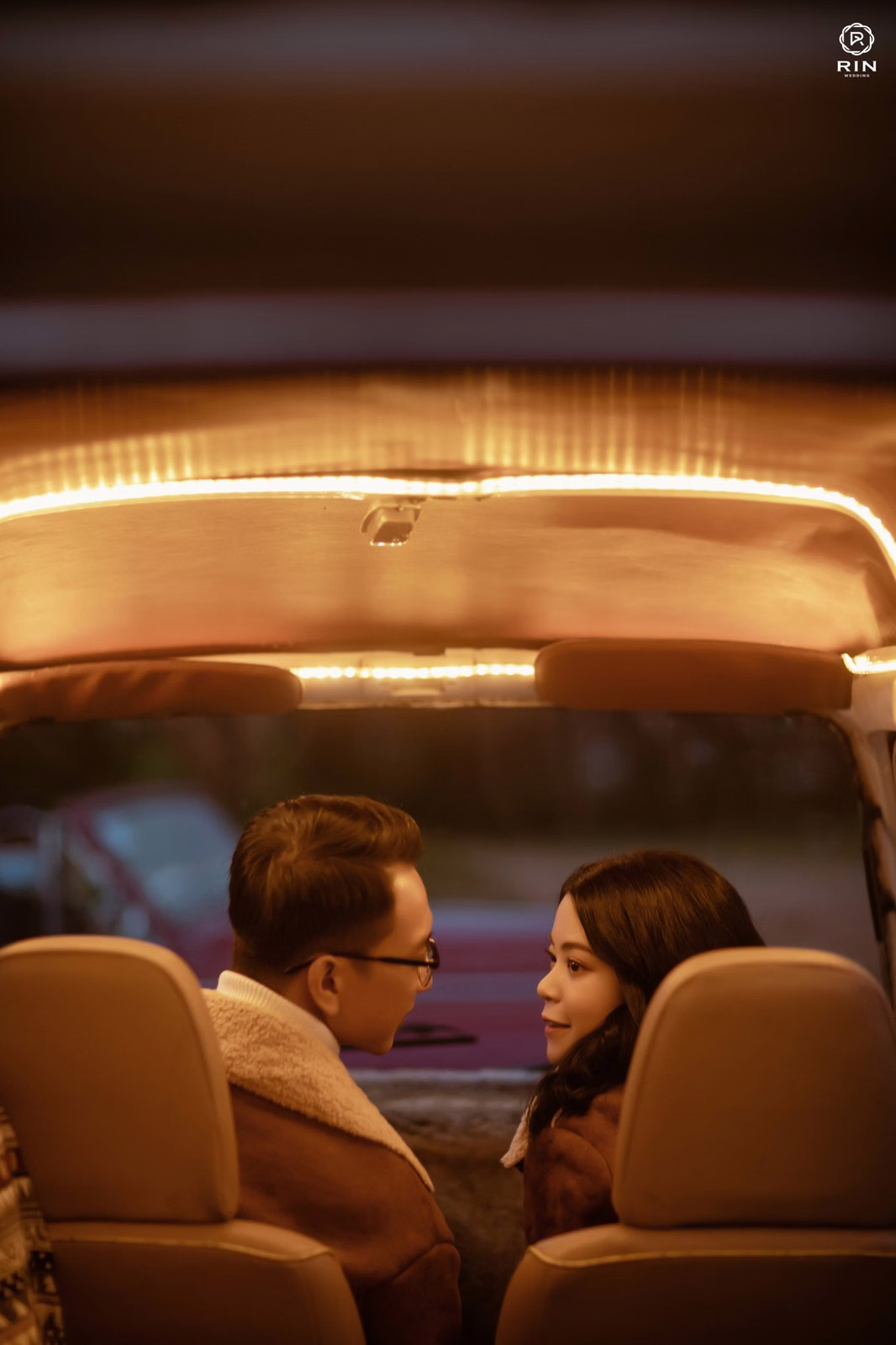 The couple sits inside a car and turns toward each other under warm yellow light.