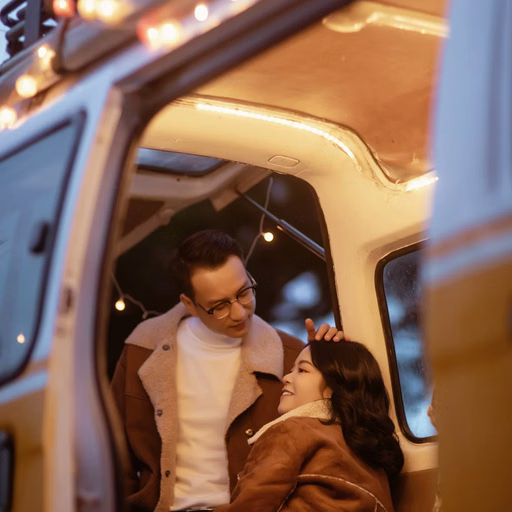 A groom looks at the bride seated in a camper beneath warm lights.