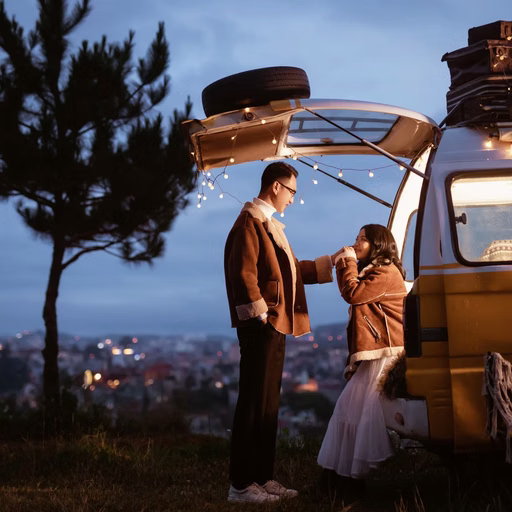 A couple chatting beside a camper vehicle, with city lights in the distance.