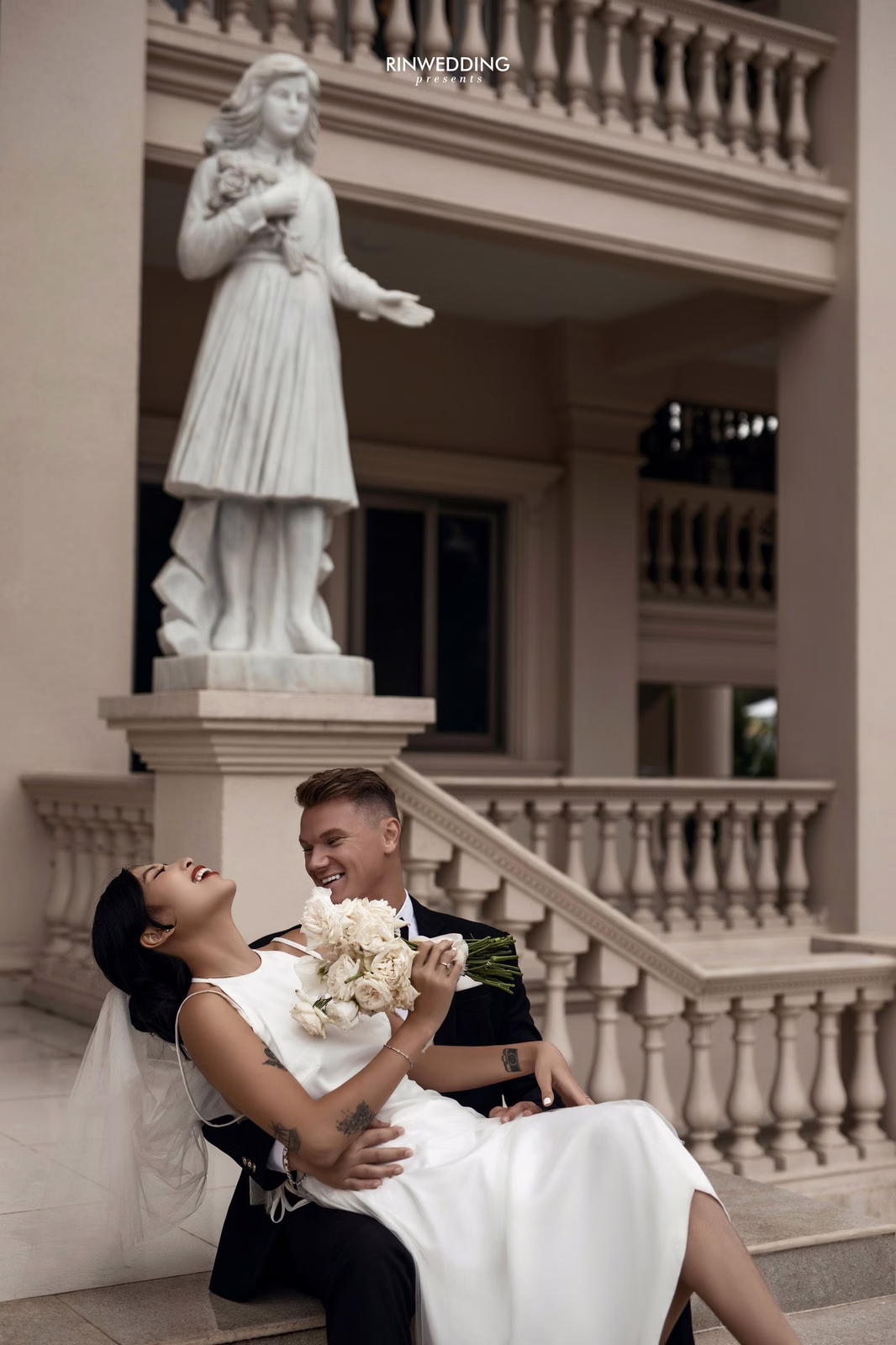 The groom supports the bride as she leans back before a statue and staircase at Don Bosco Đà Lạt.