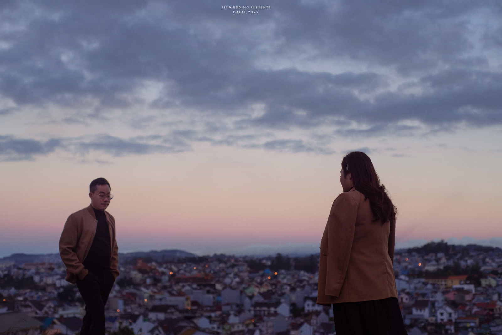 The couple looks at each other from a high viewpoint overlooking the city at dusk.