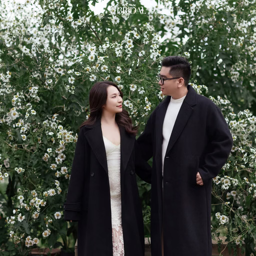 A couple in black coats in front of white flowers in a Đà Lạt café garden.