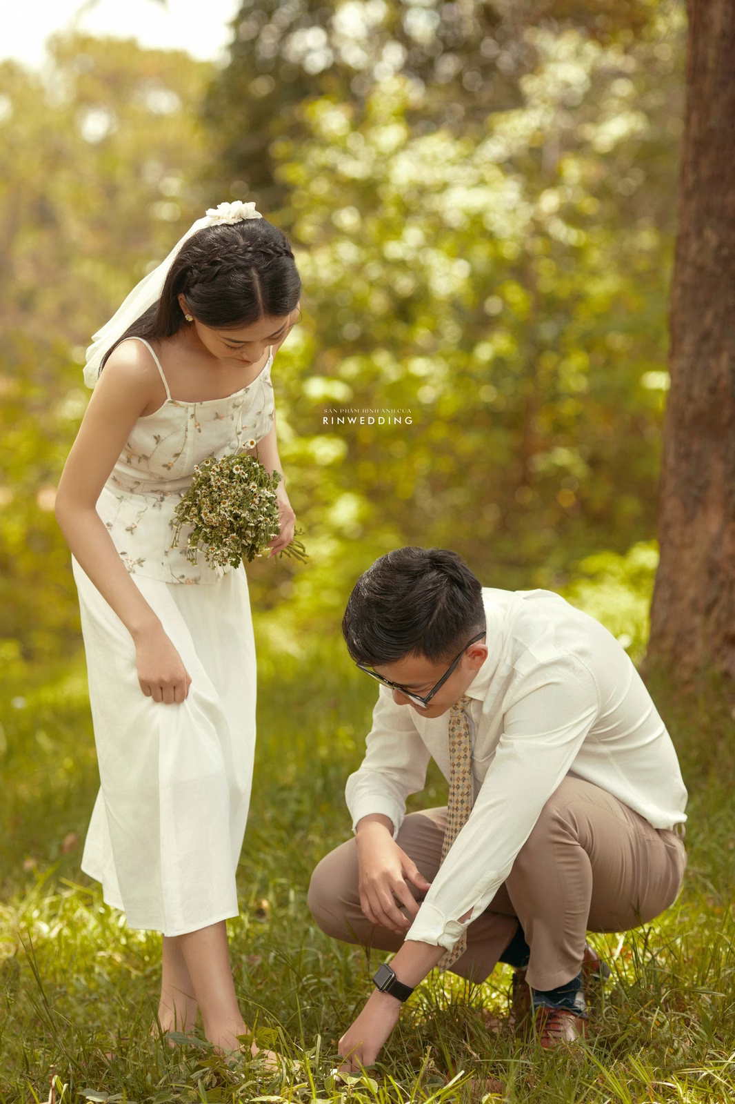 A groom bends down to adjust the bride's shoe on the lawn at Dinh Tỉnh Trưởng.