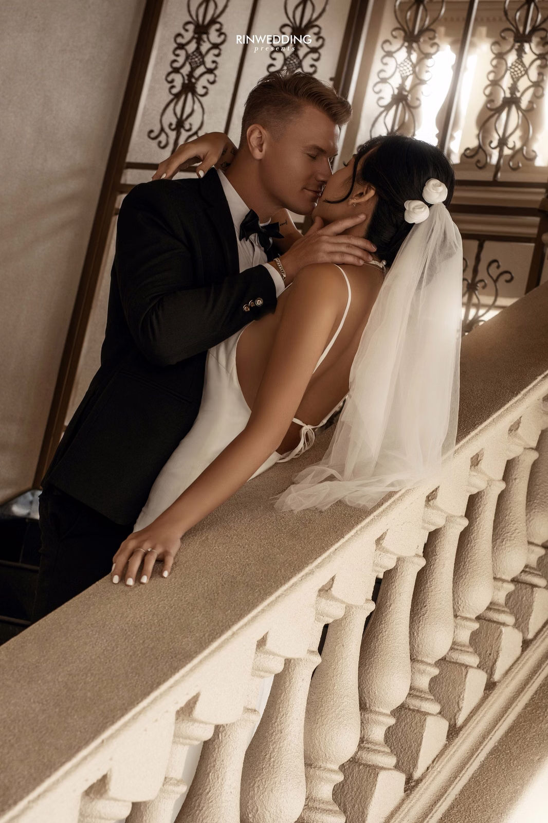 A groom kisses the bride's shoulder beside a stair railing at Don Bosco Đà Lạt.