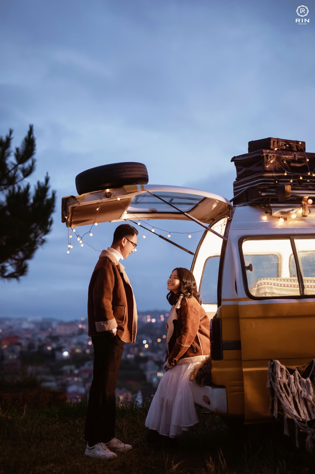 The couple stands beside the door of a camper van beneath the twilight sky.