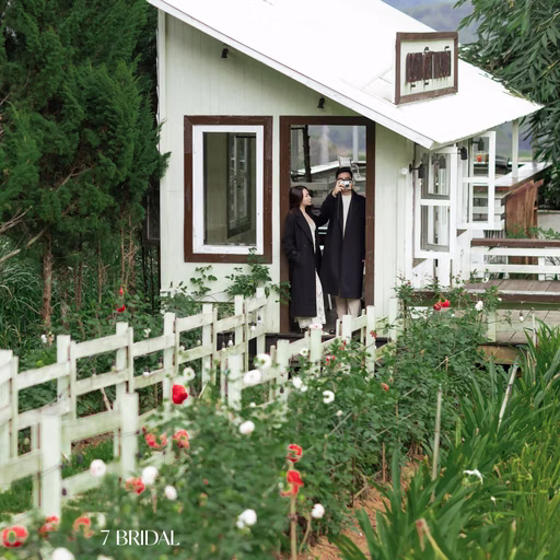 The couple stand before a white house beside a fence and flower garden at a Đà Lạt café.