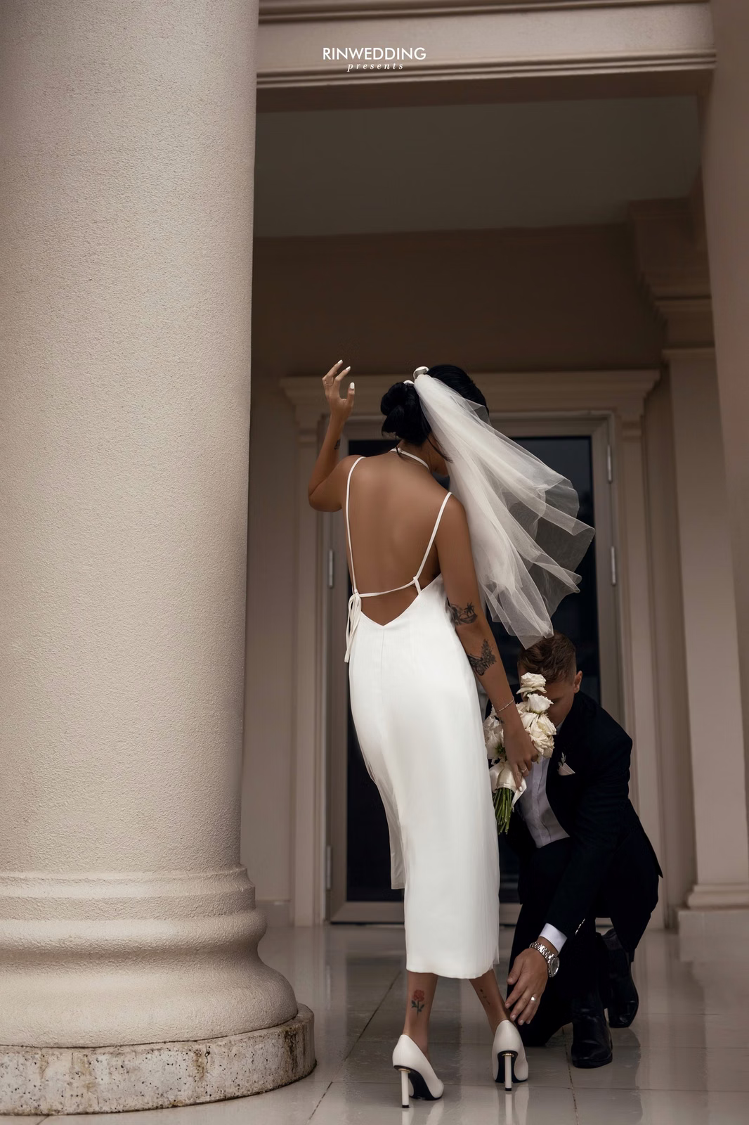 The bride poses beside a stone column with the groom in a Don Bosco corridor.