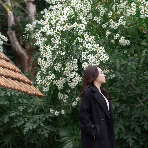 A woman in a black jacket stands beside a tiled roof and white blossoms in a Đà Lạt café.