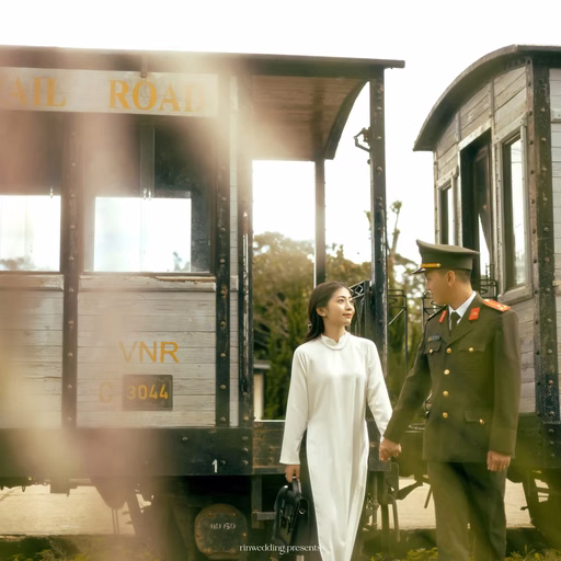 The couple stands beside a train carriage in sunshine at Ga Đà Lạt.