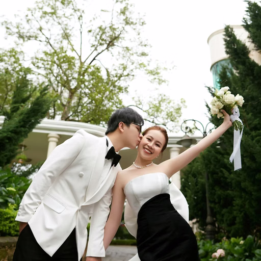 The groom kisses the bride's cheek as she raises her bouquet in a garden.