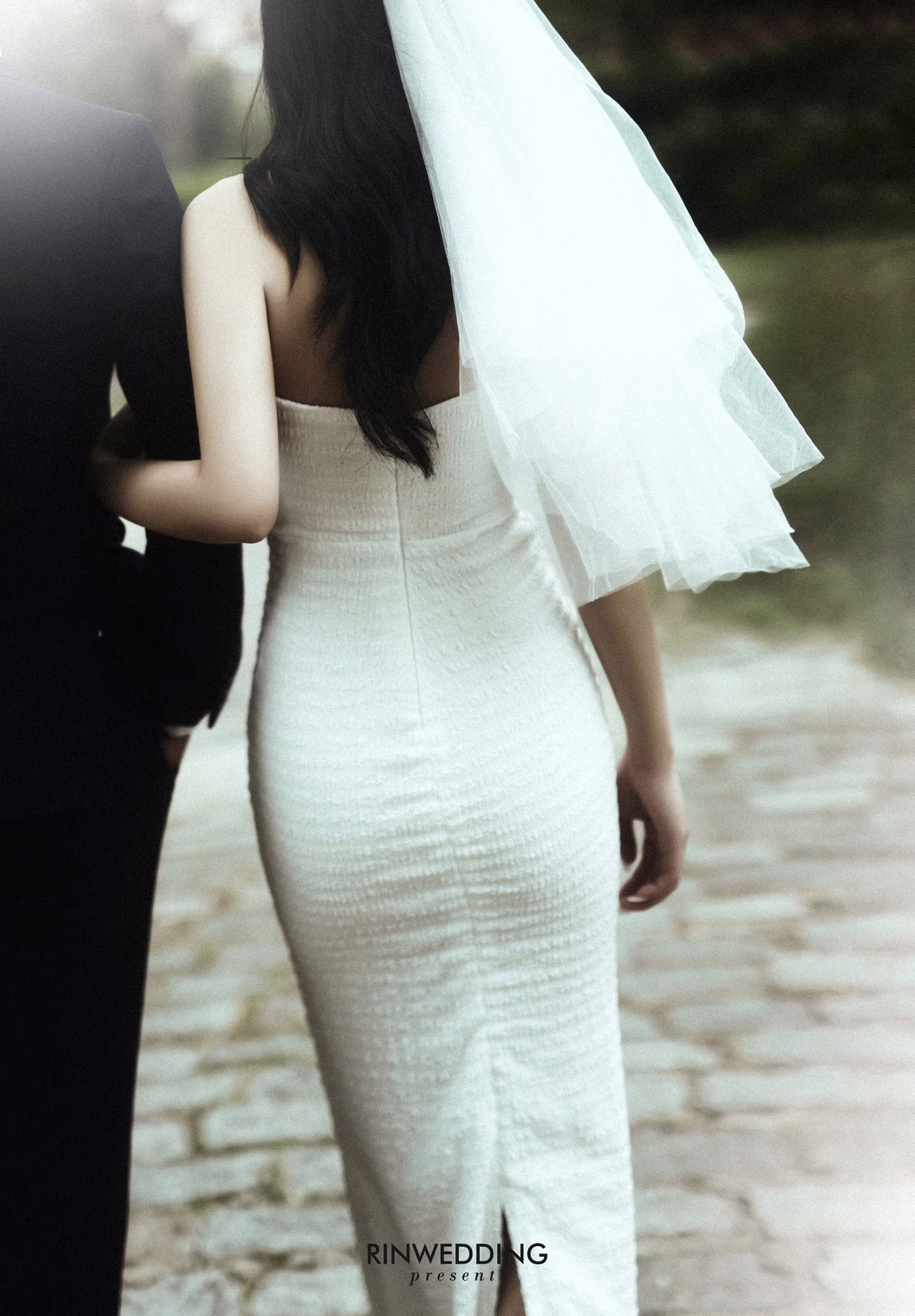 The bride in a fitted gown and veil, viewed from behind.