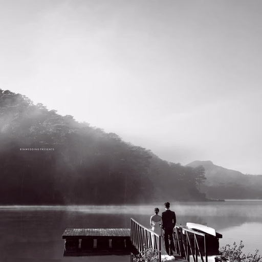 Black-and-white wedding photograph of a couple on a wooden bridge beside a misty lake.