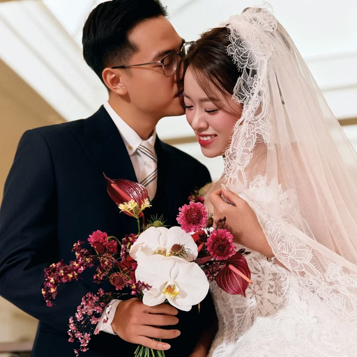 The groom kisses the forehead of the bride holding a red-and-white bouquet.