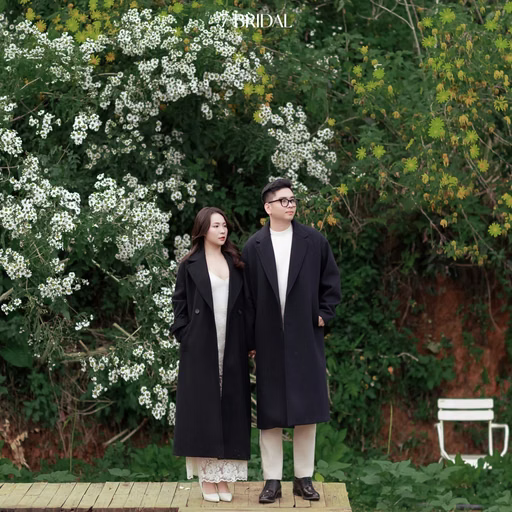 The couple in black coats stands together in a Đà Lạt café flower garden.