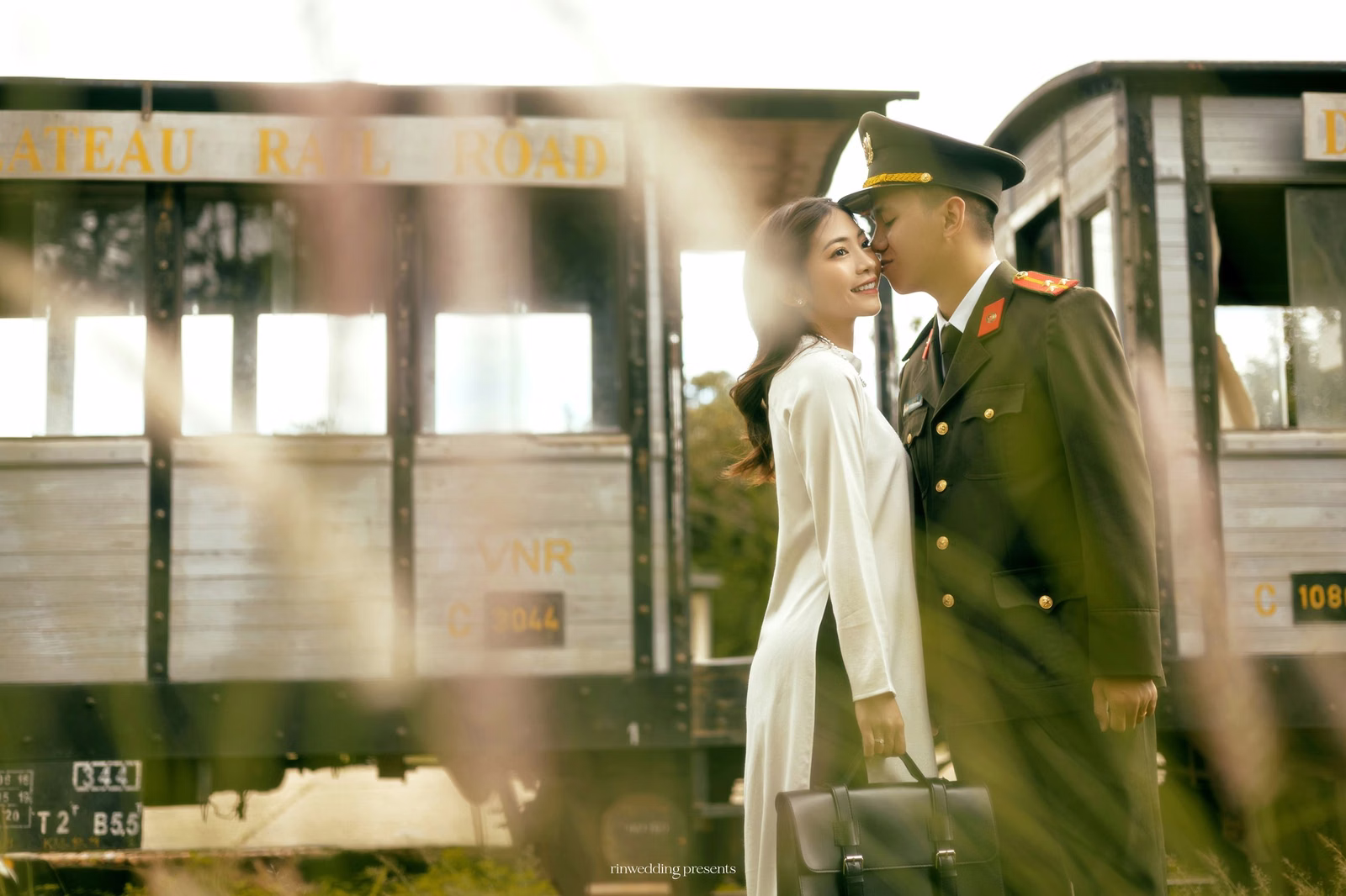 The bride in a white áo dài leans beside the groom in military uniform at Ga Đà Lạt.