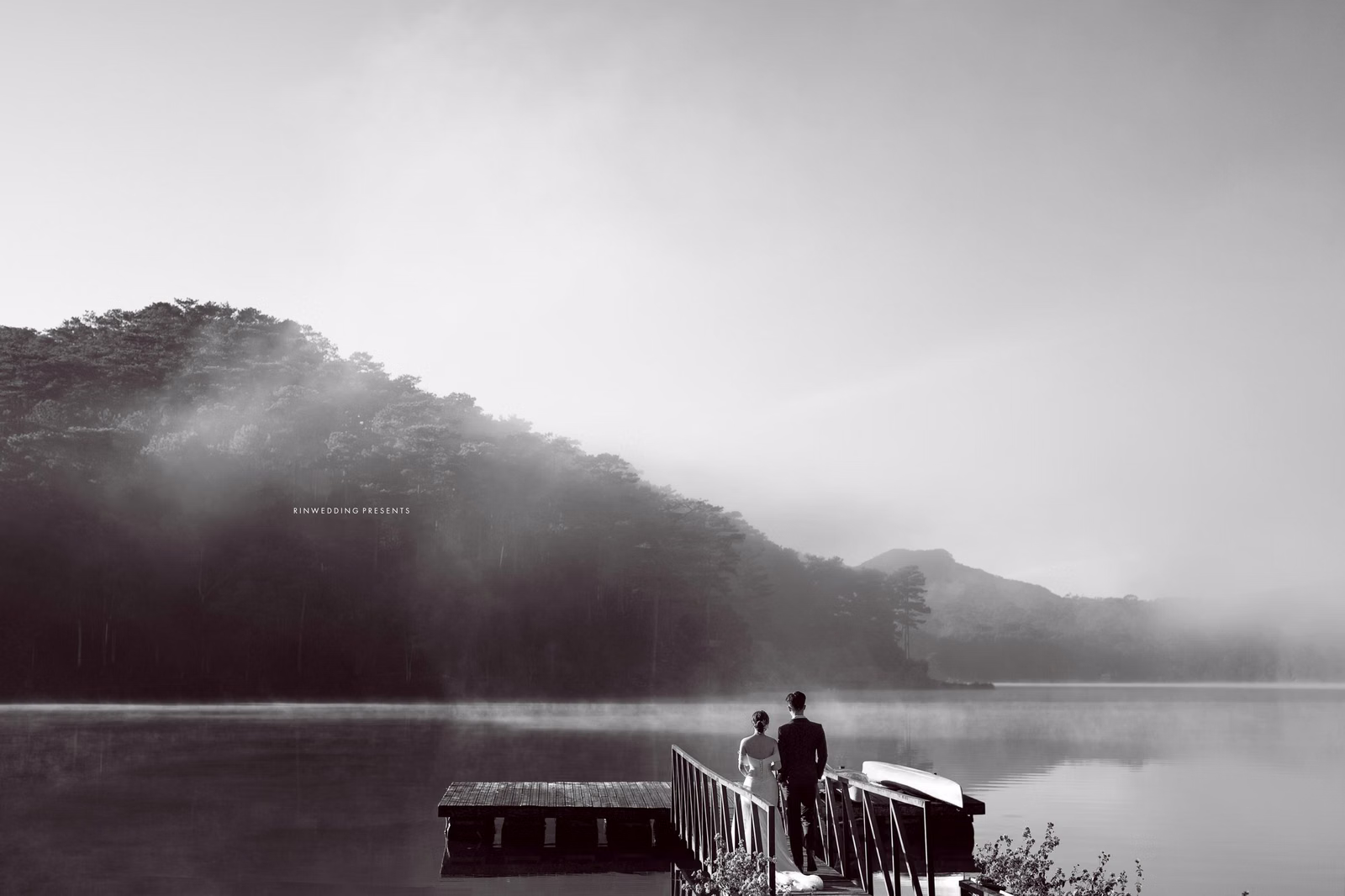 Black-and-white wedding photograph of a couple on a wooden bridge beside a misty lake.