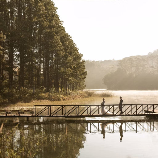The bride and groom holding hands on a wooden bridge beside a lake and pine forest.