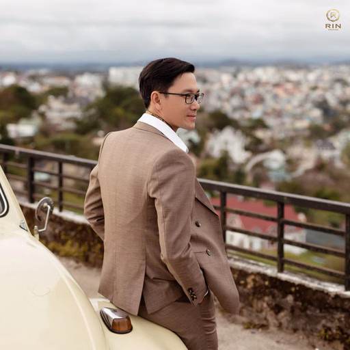 The groom in a brown suit stands beside a vintage car with Đà Lạt city behind him.