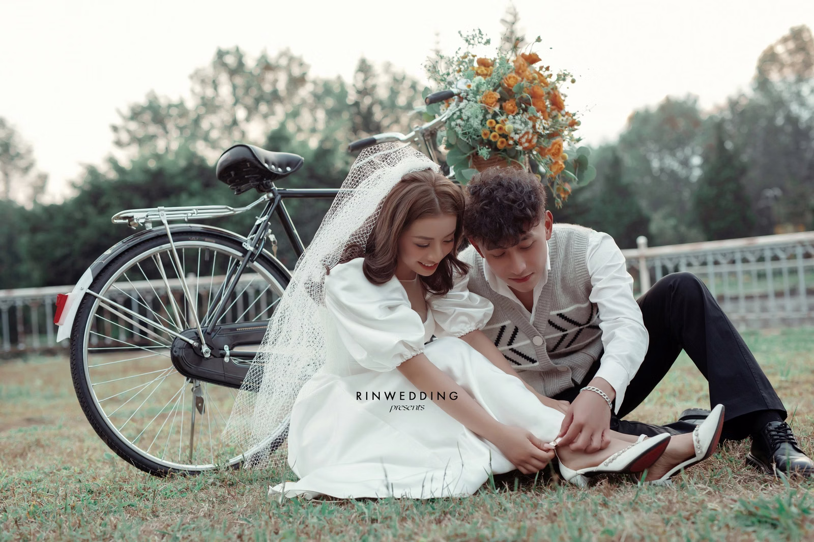 The bride and groom sit on the grass beside a bicycle and orange flowers.