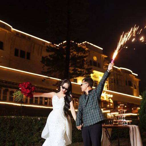 A bride and groom hold sparklers before a hotel at night.