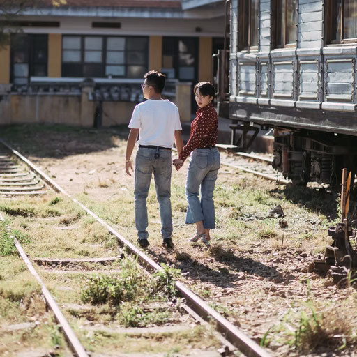 The couple holds hands walking beside a carriage and tracks at Ga Đà Lạt.