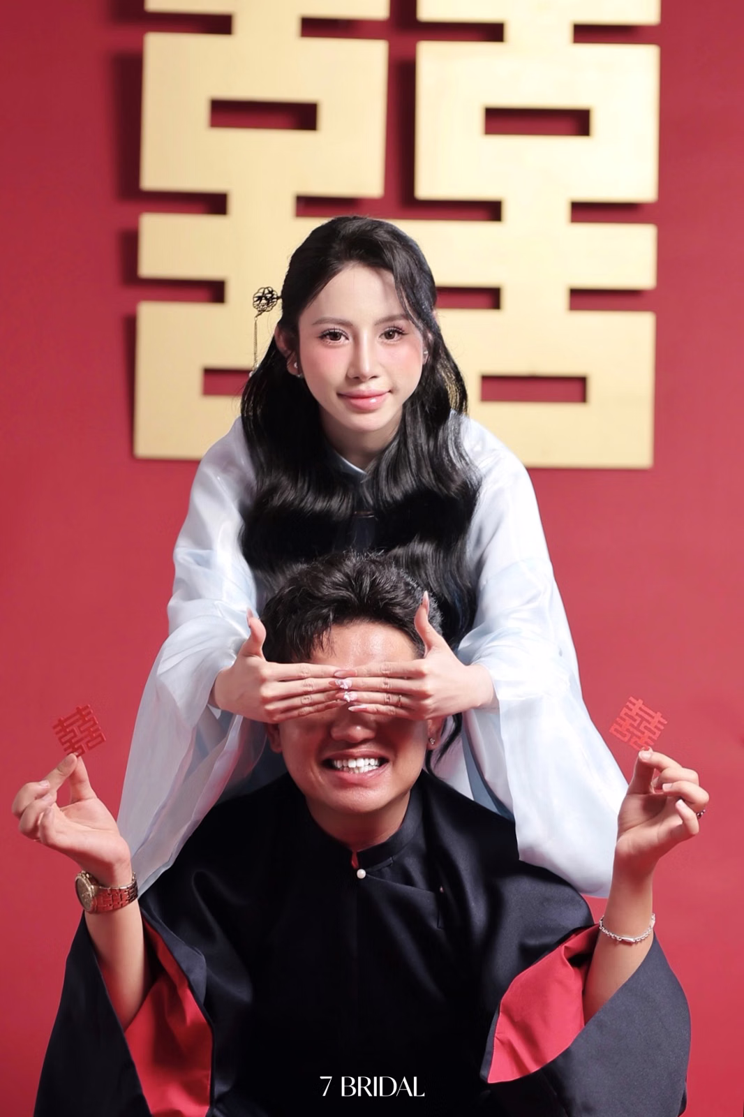 The bride playfully covering the groom’s eyes against a red backdrop.