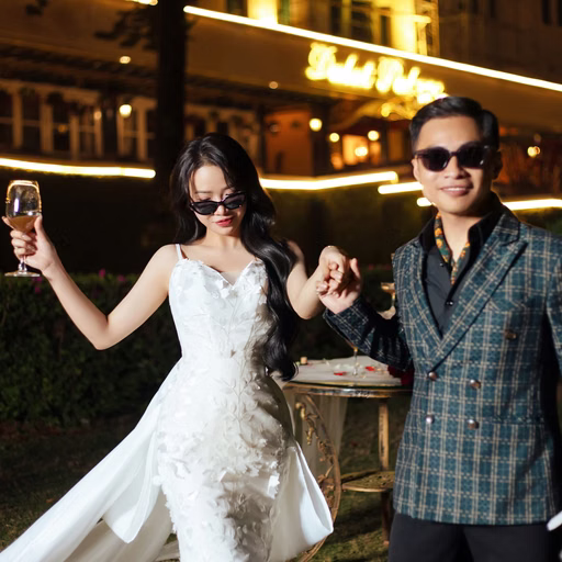 The bride and groom wear sunglasses and raise glasses outside a hotel at night.