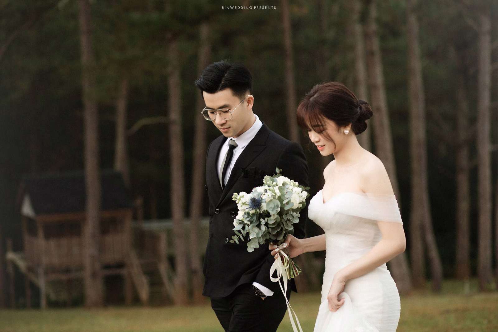 The bride and groom walk together with a white bouquet beside the pine forest.