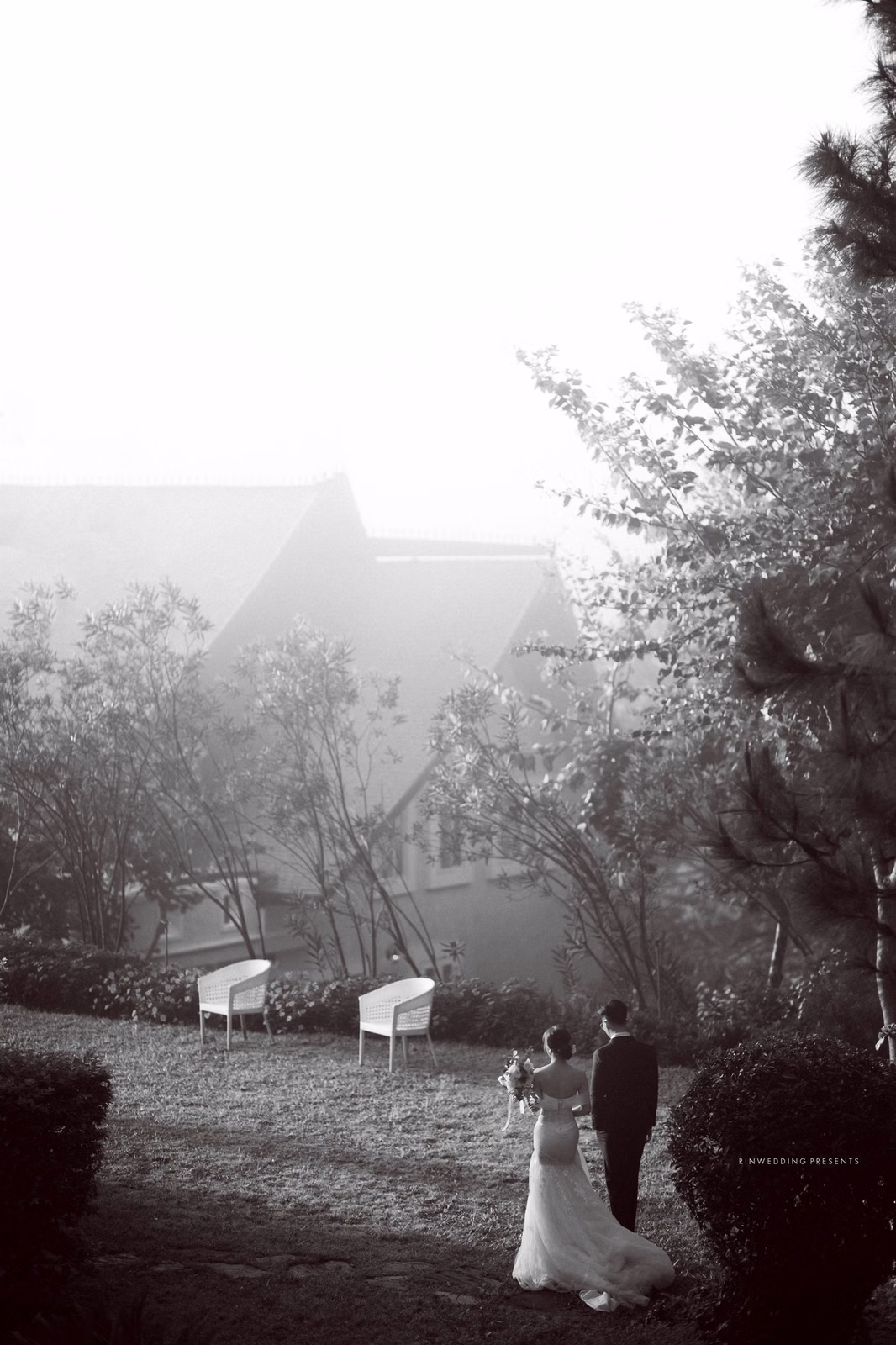 Black-and-white wedding photograph of the bride and groom before a villa with a pitched roof.