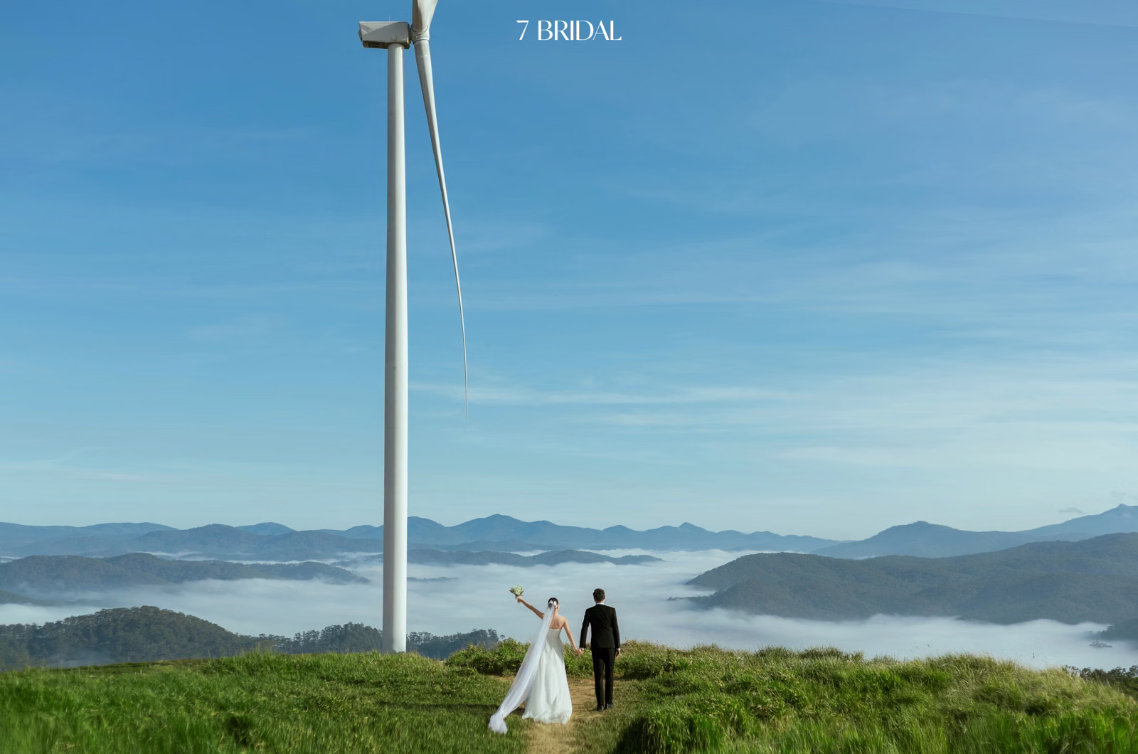 The couple stand beneath a wind turbine blade, looking out over a sea of clouds at the Cầu Đất wind farm