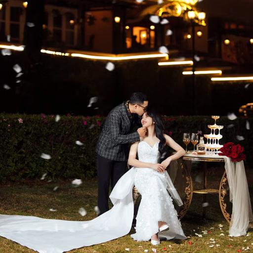 The groom kisses the bride seated beside a reception table outside a hotel at night.
