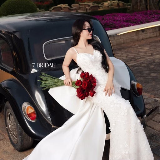 The bride wears sunglasses and leans beside a black vintage car.
