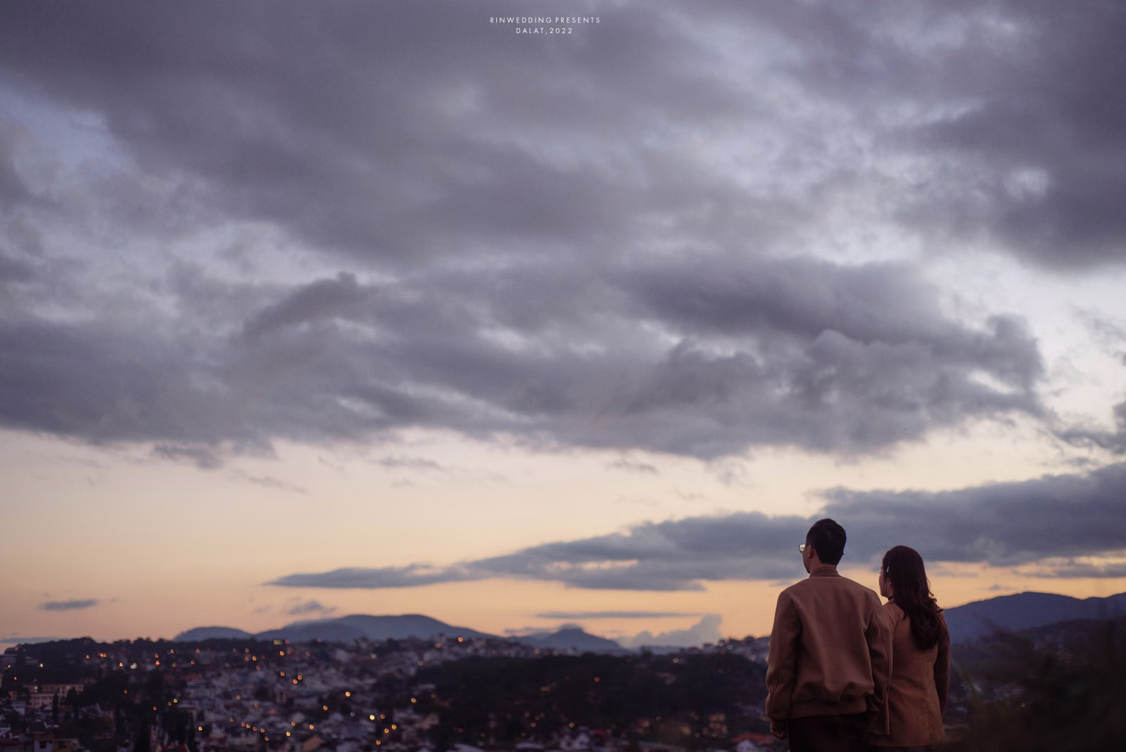 A couple looking out over Đà Lạt under a cloudy evening sky.
