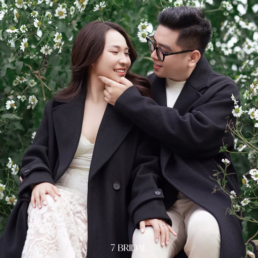 A man touching his girlfriend's chin as they sit before a white floral trellis.